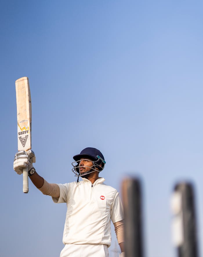 Cricketer in white uniform holding a bat under the clear sky, celebrating success on the field.